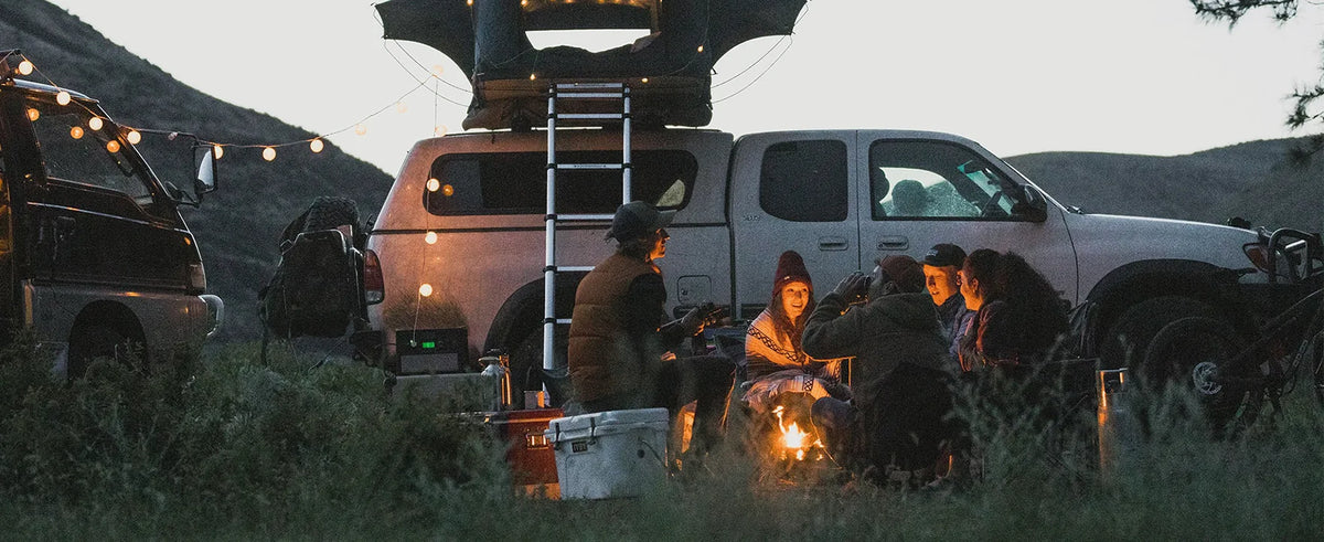 Friends around a campfire next to an overland truck with a rooftop tent at dusk
