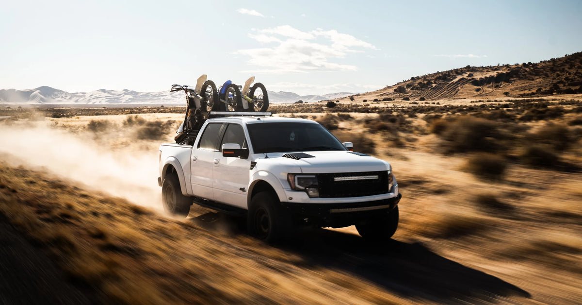 White pickup truck with three dirt bikes on a Big Days Chase Rack, driving a dusty desert track