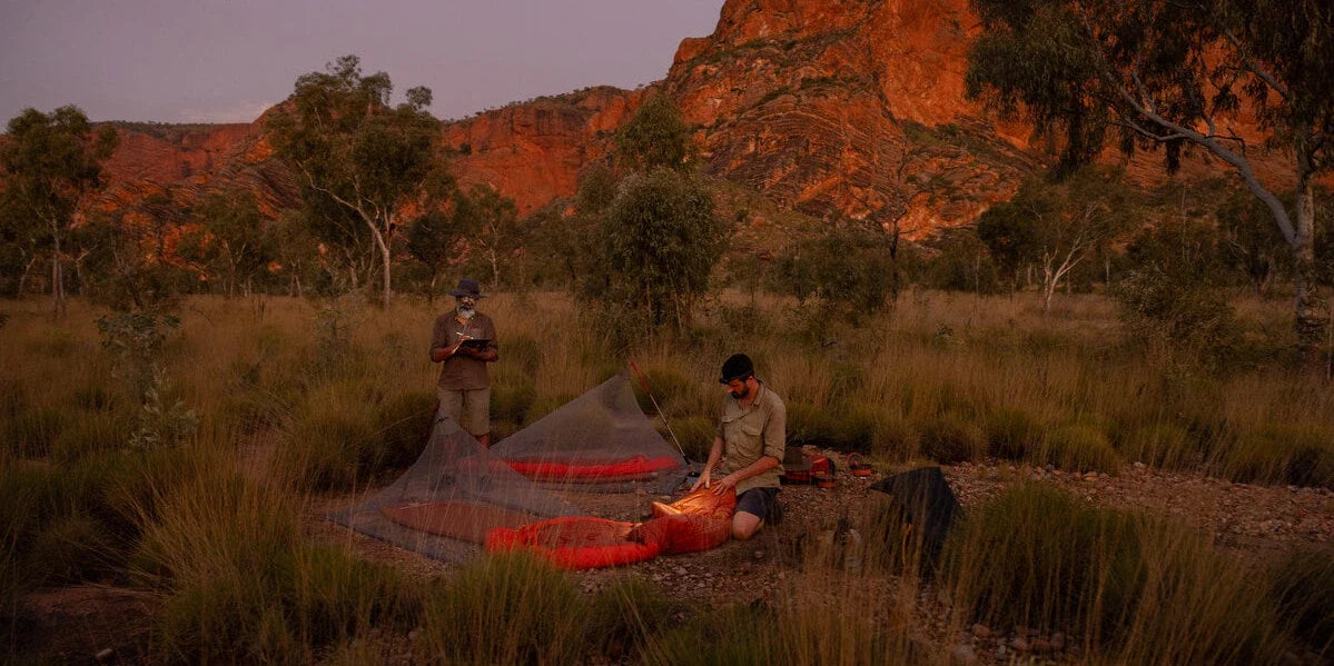 Two campers setting out red Sea to Summit sleeping bags under mesh shelters below red rock cliffs at dusk