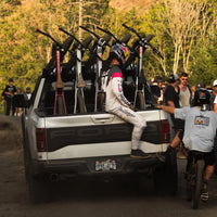 People with bicycles in a truck bed on a dirt road with trees in the background