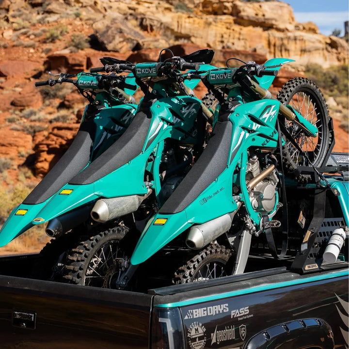 Three teal dirt bikes upright on a Big Days Chase Rack in a black pickup bed with red rock cliffs behind