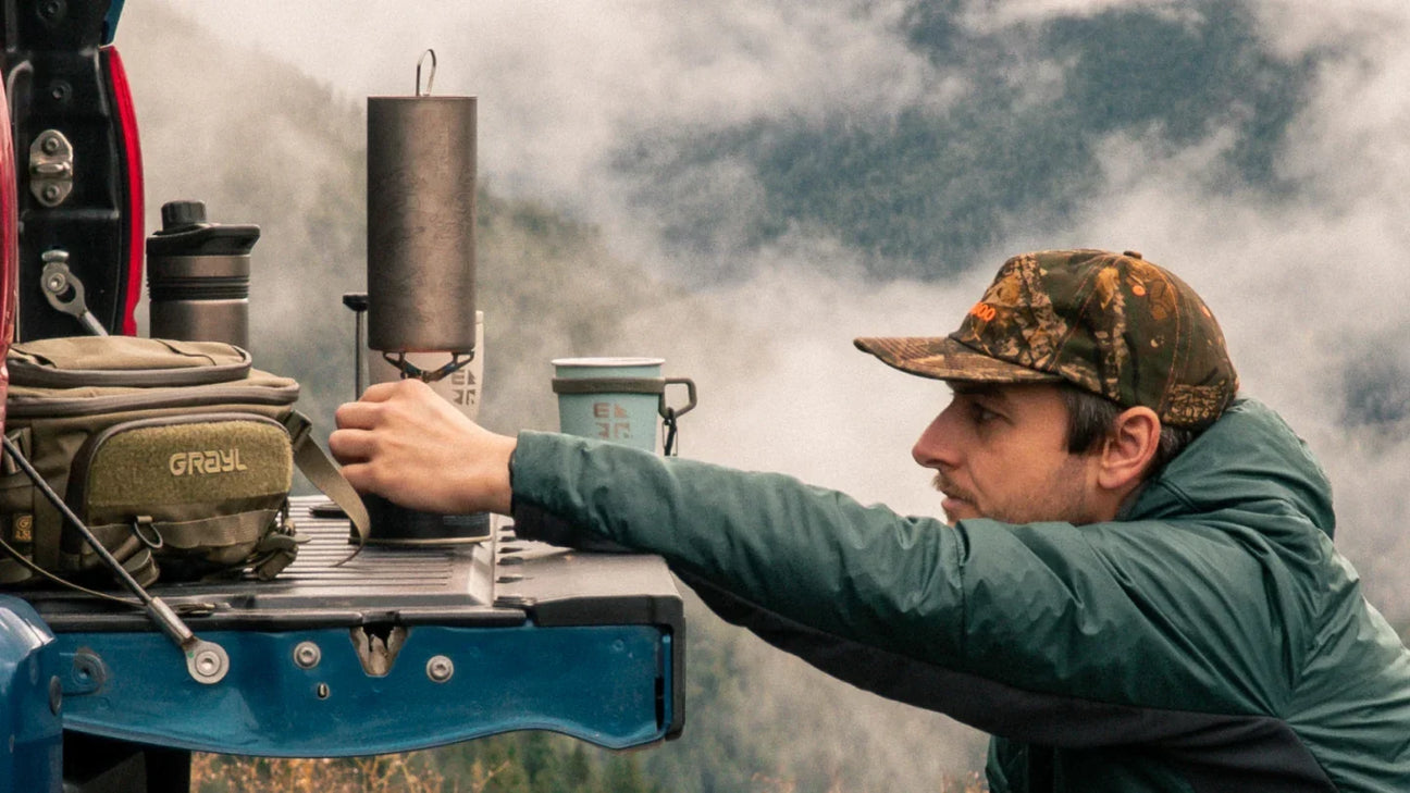 GRAYL GeoPress Ti titanium cup heating water on a camp stove set on a truck tailgate, with a GRAYL pack and misty mountains behind