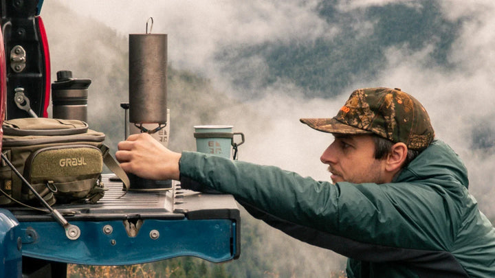 GRAYL GeoPress Ti titanium cup heating water on a camp stove set on a truck tailgate, with a GRAYL pack and misty mountains behind
