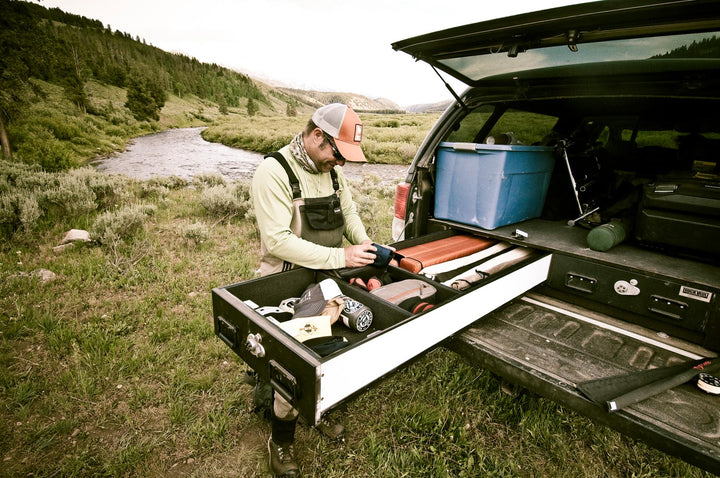 Angler in waders packing fly fishing gear from a slide-out truck bed drawer beside a mountain river