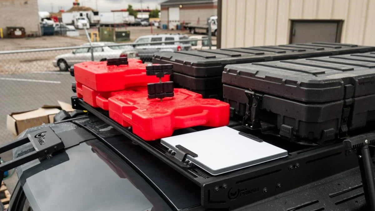 Prinsu roof rack on a dark SUV carrying red fuel cans, two black storage cases and a flat white panel