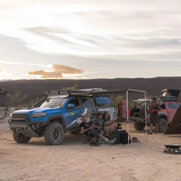 Blue Toyota Tacoma with a loaded Prinsu roof rack and awning set up at a desert camp at sunset, friends relaxing in camp chairs