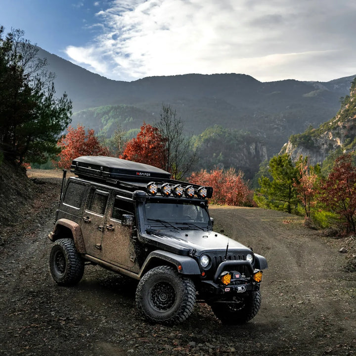 Muddy Jeep with a loaded roof rack and light bar parked on a dirt road in autumn mountains
