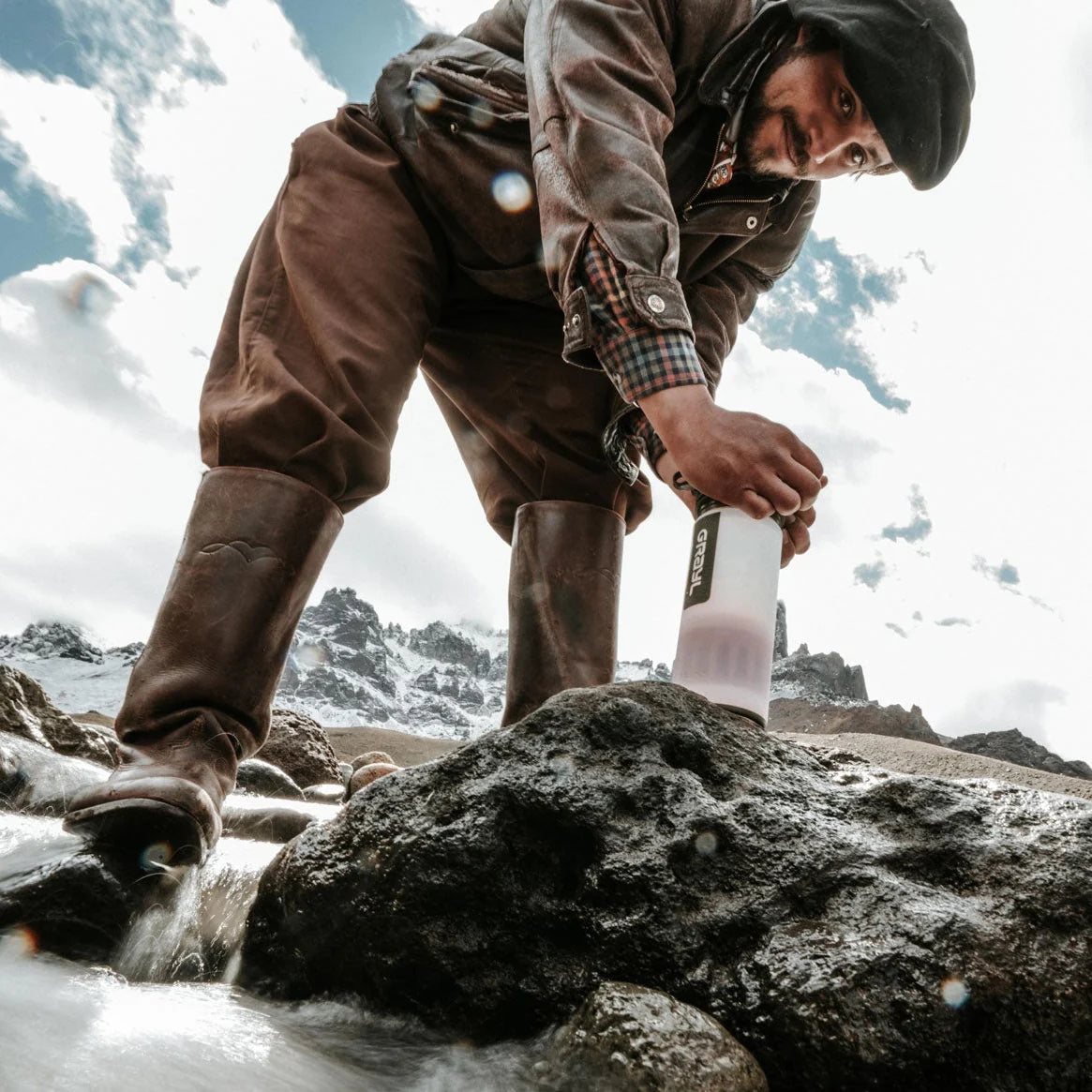 Man pressing a GRAYL water purifier bottle on a rock beside a mountain stream