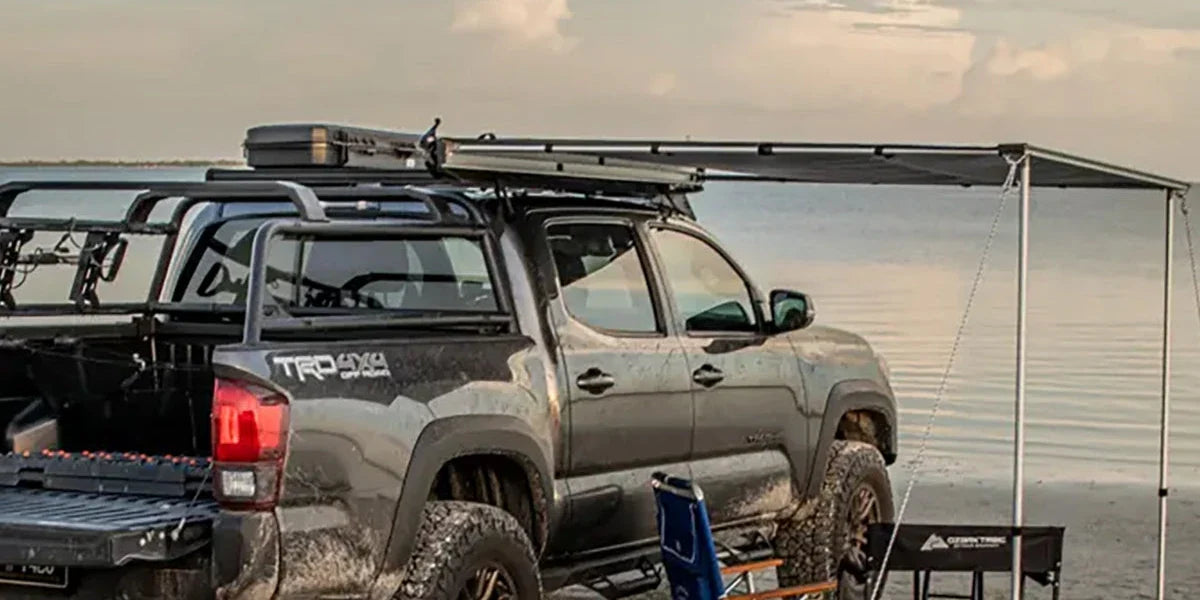 Toyota Tacoma with a Body Armor 4x4 awning set up over a lakeside camp with a chair and table
