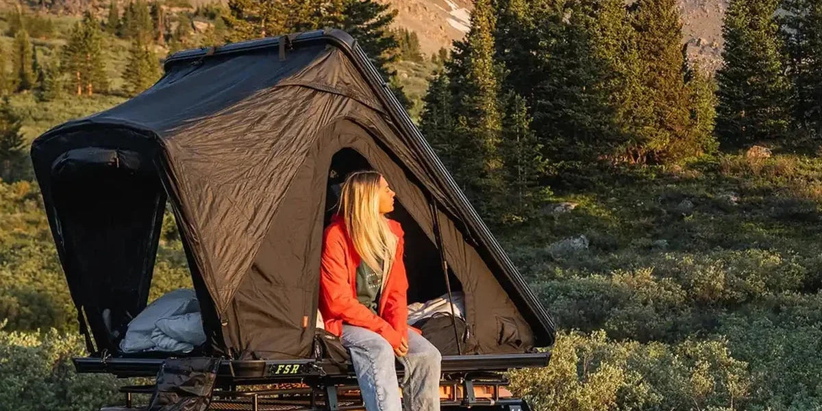 Camper sitting in the doorway of a Free Spirit Recreation rooftop tent in a pine forest