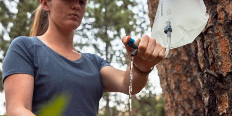 Camper filling a cook pot from a hanging HydraPak water bag at a forest camp