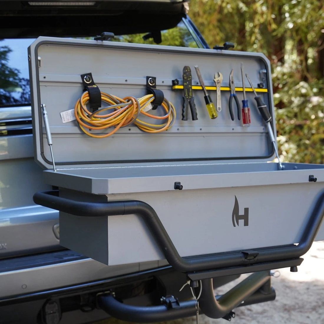Tool box attached to a vehicle with tools and cables inside, outdoors.