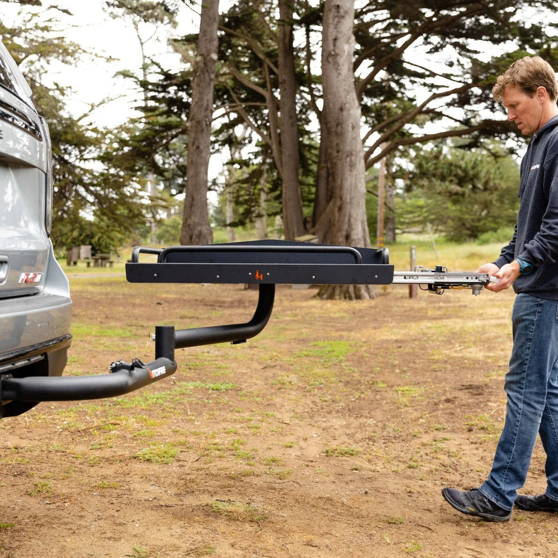 Man attaching a hitch to a car in an outdoor setting with trees in the background