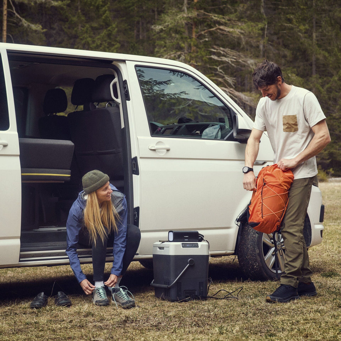 Two people preparing for a trip by a van with a cooler and camping gear.