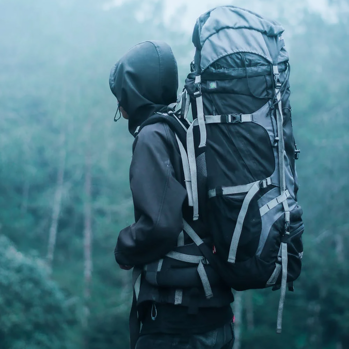 Hiker wearing a hooded jacket and carrying a large black trekking backpack in a misty forest.