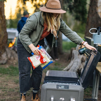 Woman using portable camping fridge at outdoor campsite with fire in background