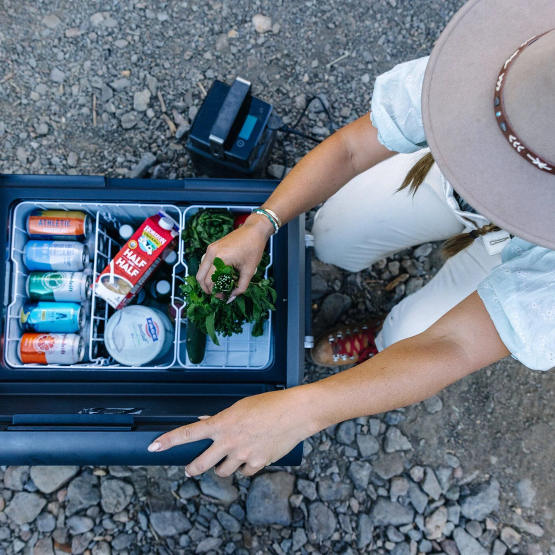 Camper opening portable fridge with drinks, greens, and half-and-half outdoors on rocky ground