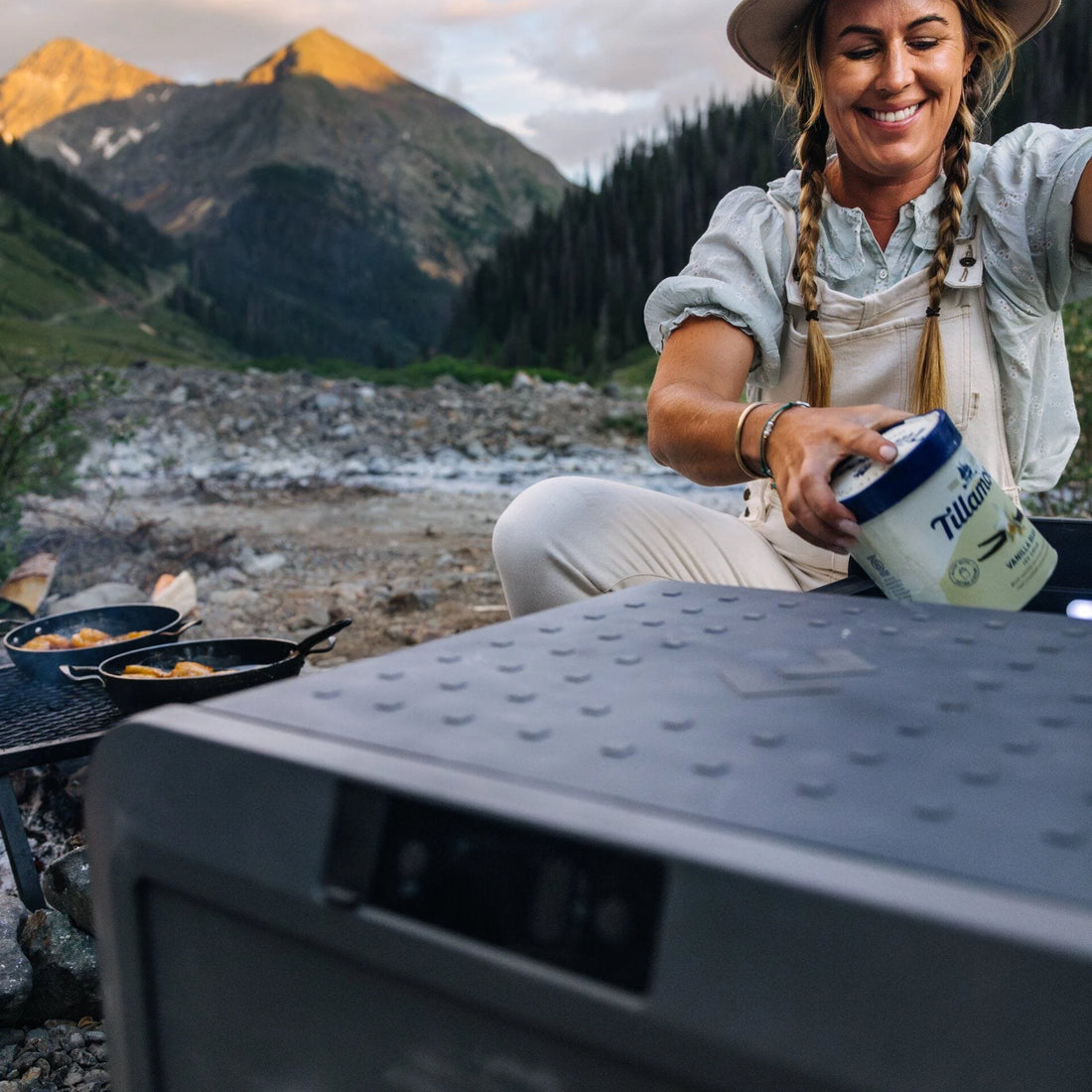 Woman camping outdoors, opening cooler with Tillamook ice cream, mountains in background