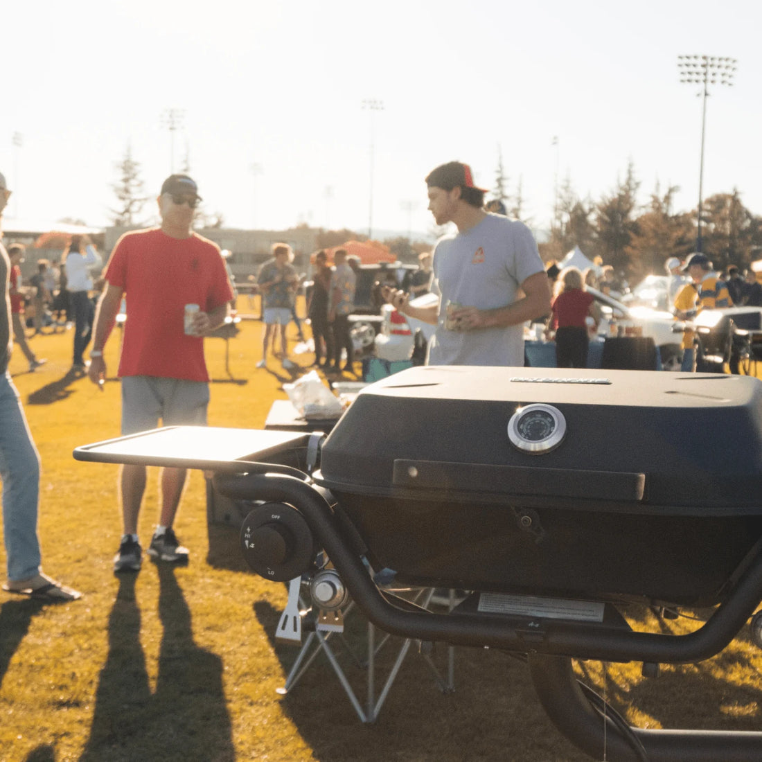 People standing around a grill at an outdoor event with cars and people in the background.