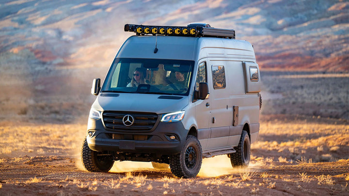 A Mercedes-Benz Sprinter camper van with a roof-mounted LED light bar drives through dusty terrain in a desert landscape at sunset.