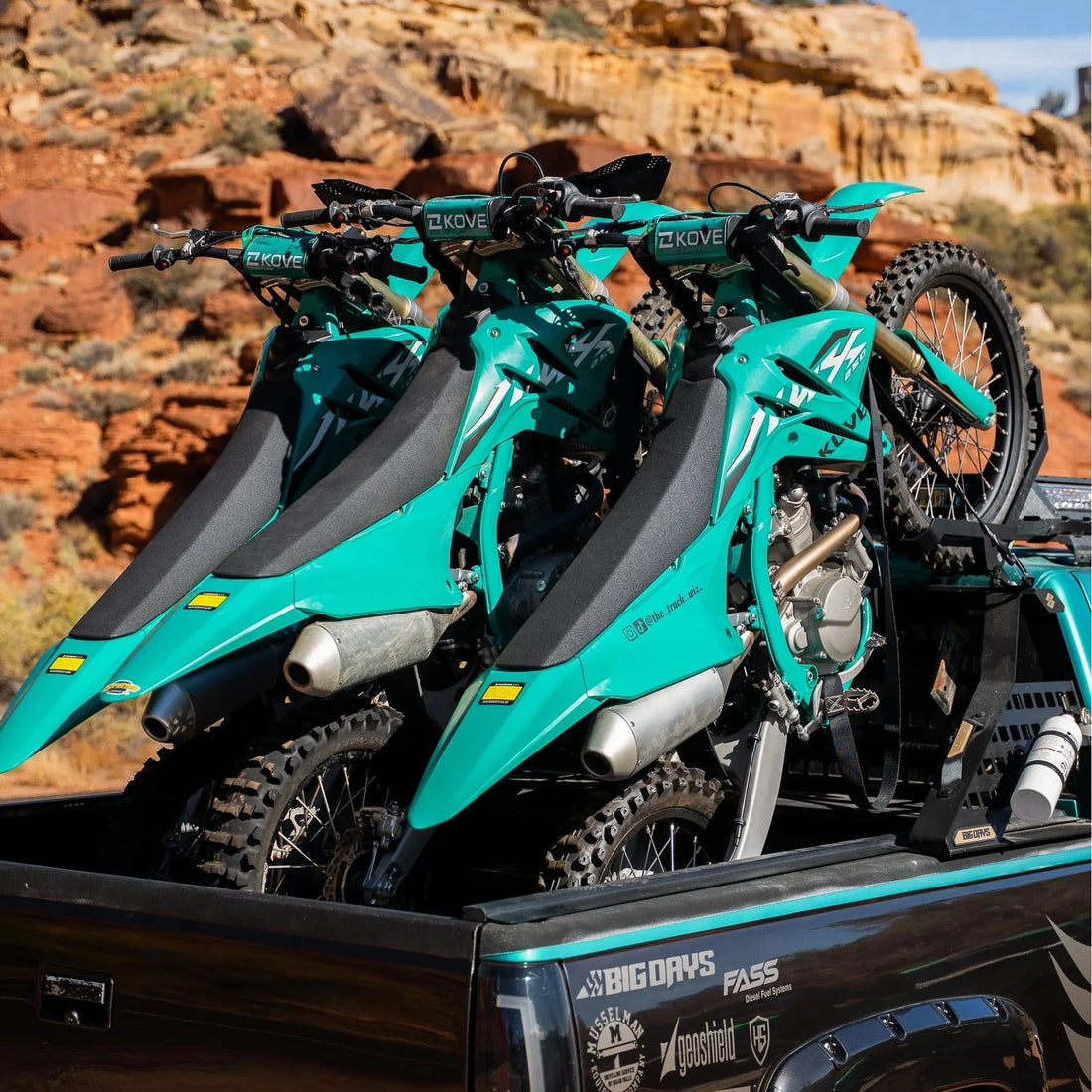 Two teal dirt bikes in the back of a pickup truck with a desert landscape in the background