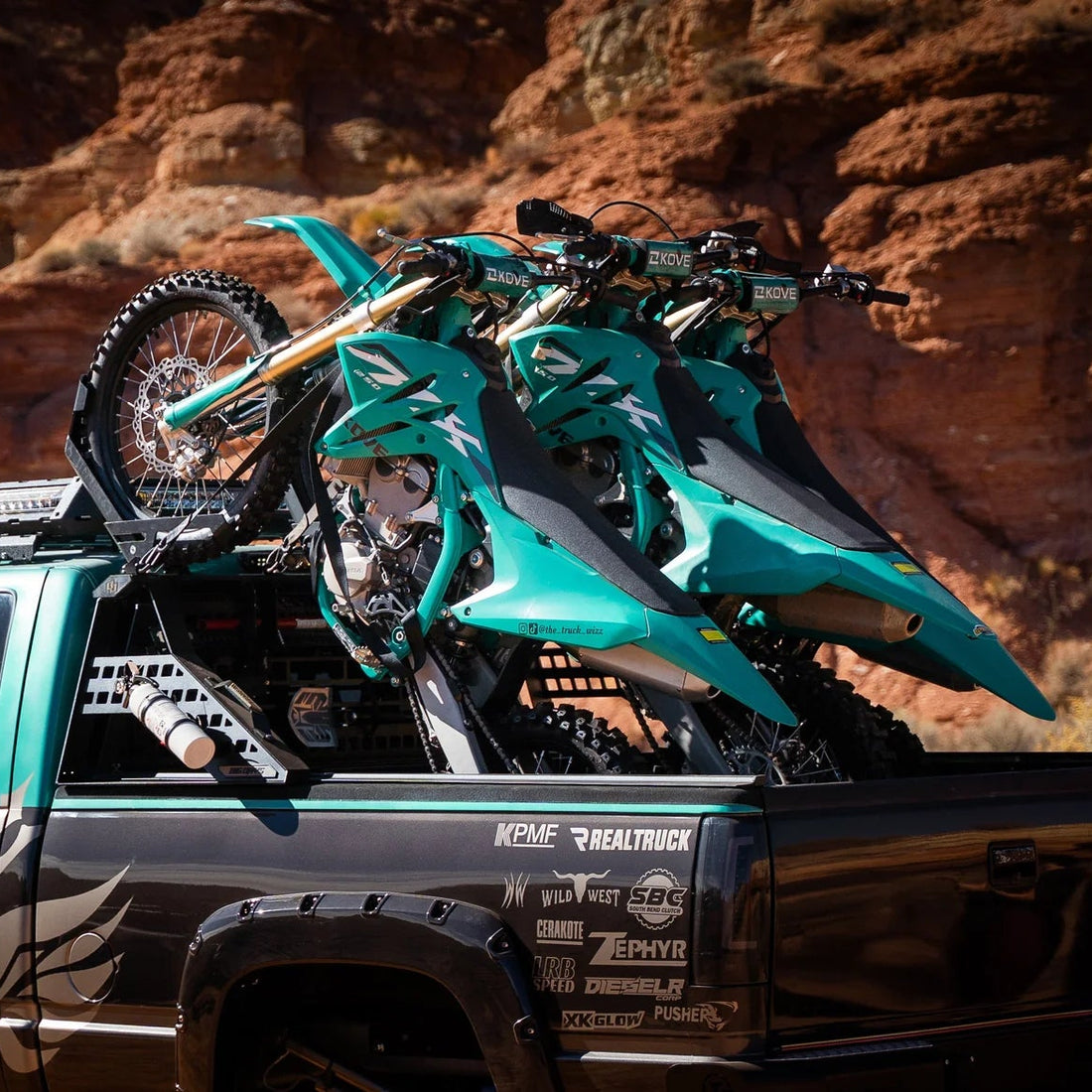 Three green dirt bikes on a black truck bed with a desert landscape in the background