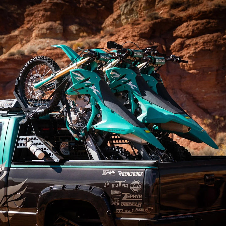 Three green dirt bikes on a black truck bed with a desert landscape in the background