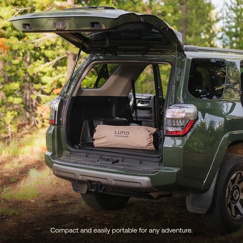 Green SUV with open trunk showing a camping setup in a forest setting, featuring the brand &
