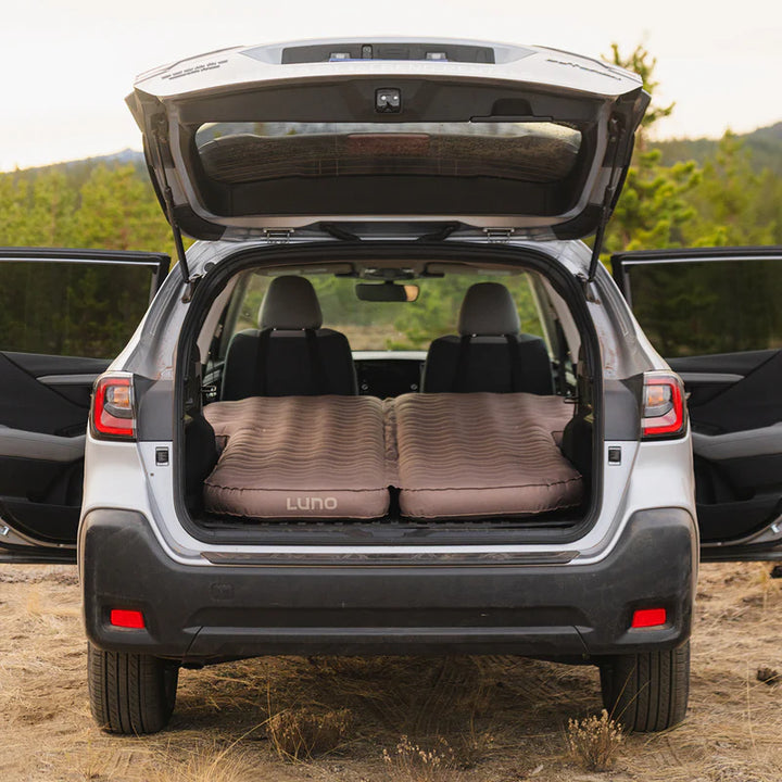 Car with open trunk showing a camping mattress branded 'Luno' in a natural setting.