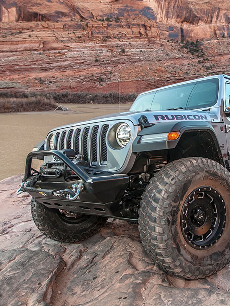 Silver Jeep Rubicon with off-road bumper and mud-terrain tires on red rocks