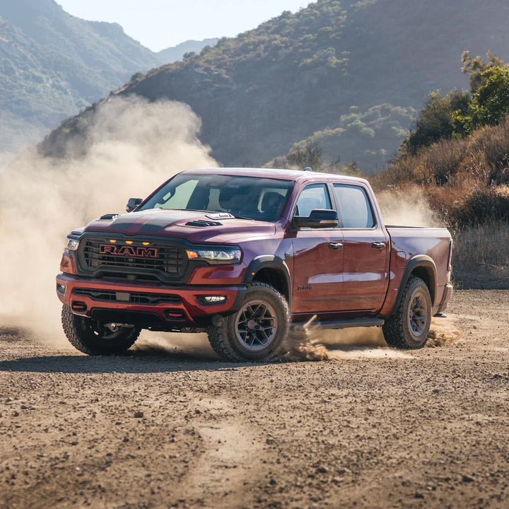 A red Ram pickup truck driving on a dusty dirt road with mountains in the background.