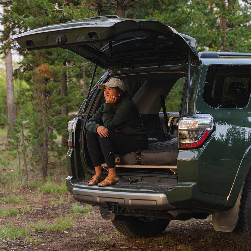 Person sitting in the open trunk of a green SUV in a forest setting