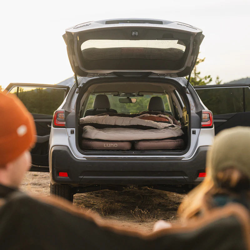 Car with open trunk showing camping setup, surrounded by people in an outdoor setting.