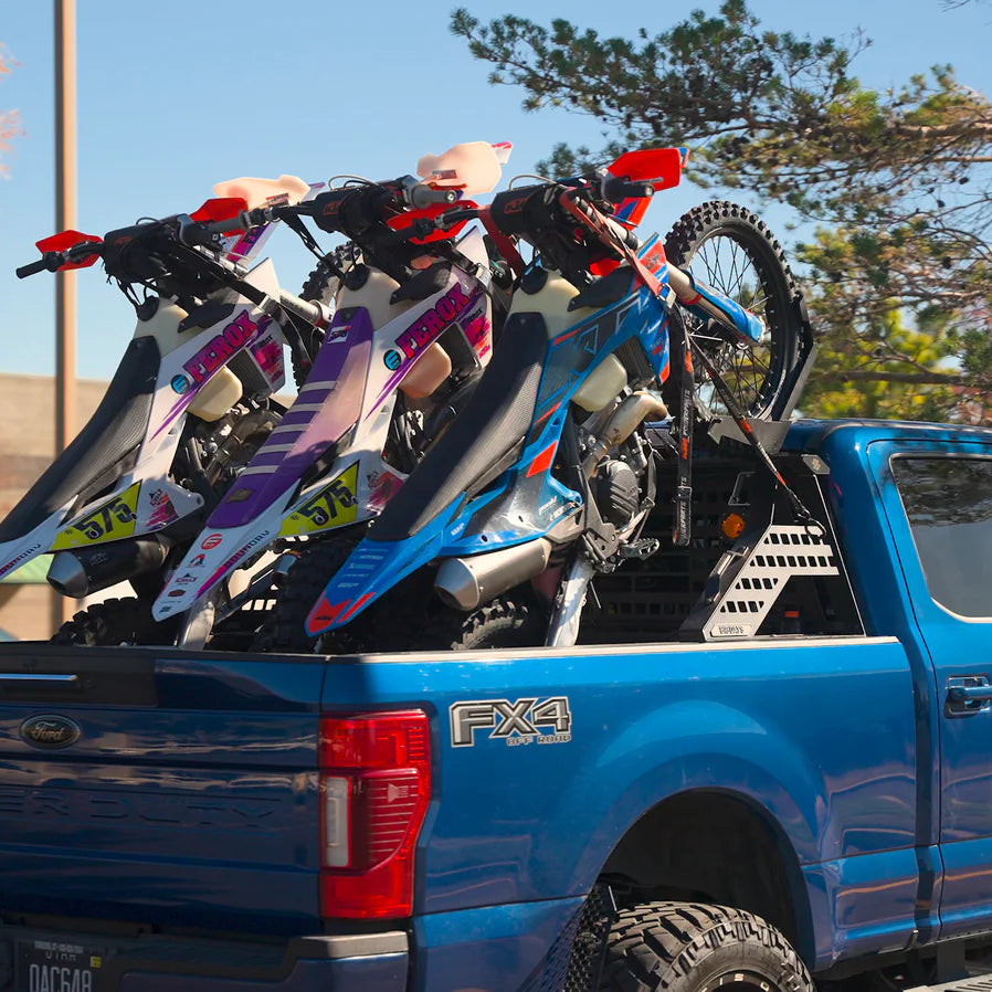 Three dirt bikes securely mounted on a blue pickup truck bed rack outdoors.
