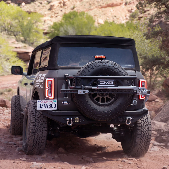 A black Bronco Spare Tire Guard mounted on the rear of a vehicle with large off-road tires, in a rugged outdoor setting.