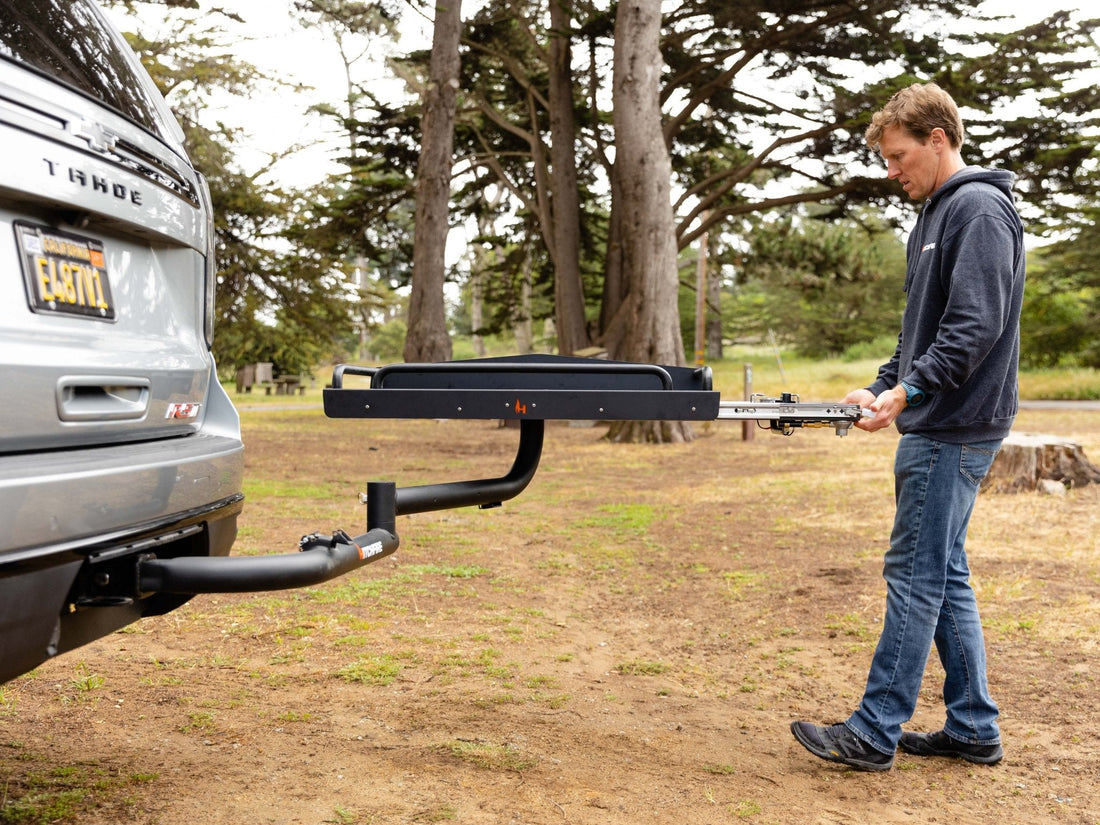 Man attaching a hitch to a car in an outdoor setting with trees in the background