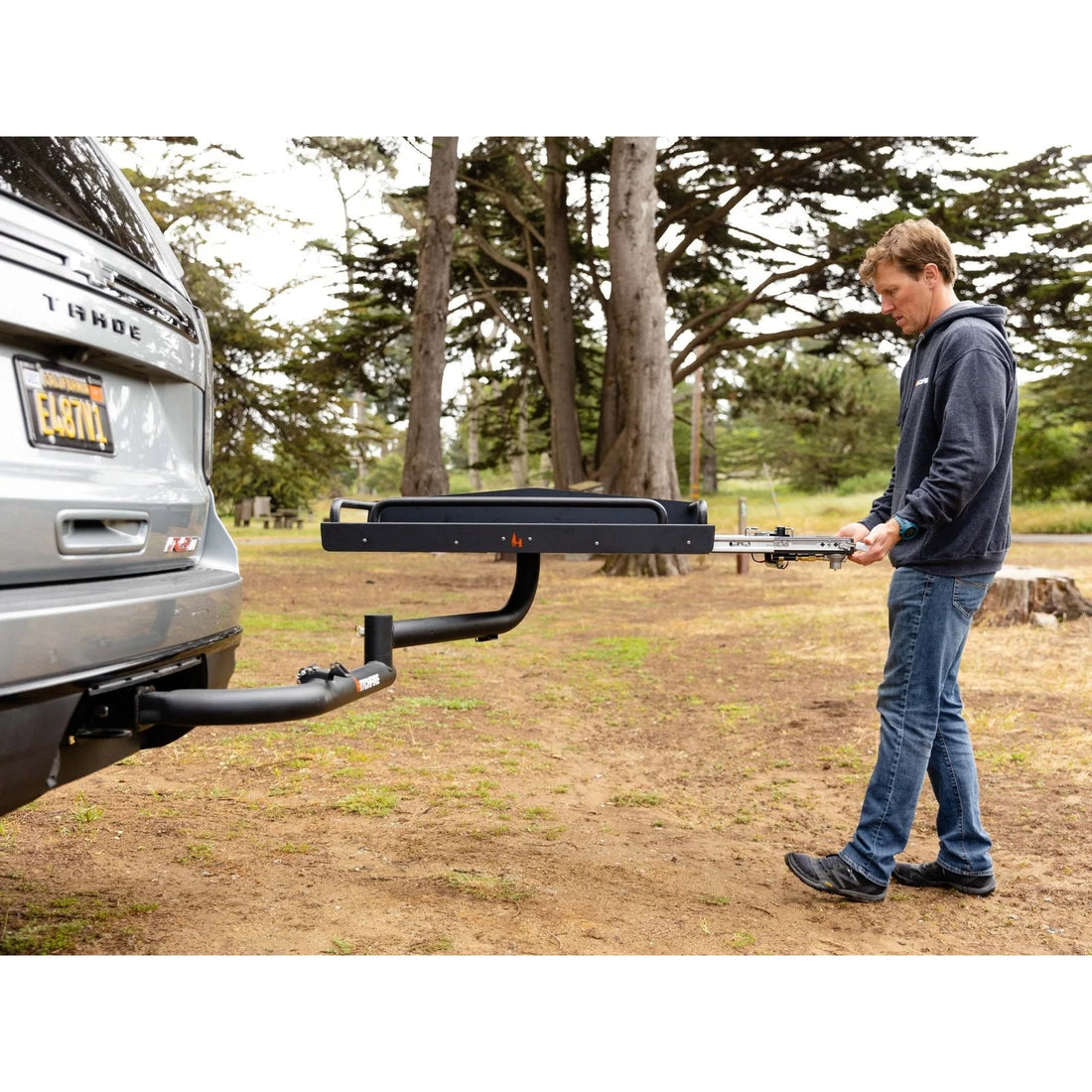 Man attaching a hitch to a car in an outdoor setting with trees in the background
