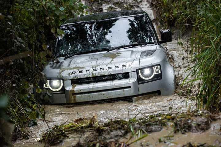 A silver Land Rover Defender driving through muddy water on an off-road trail surrounded by dense green foliage.