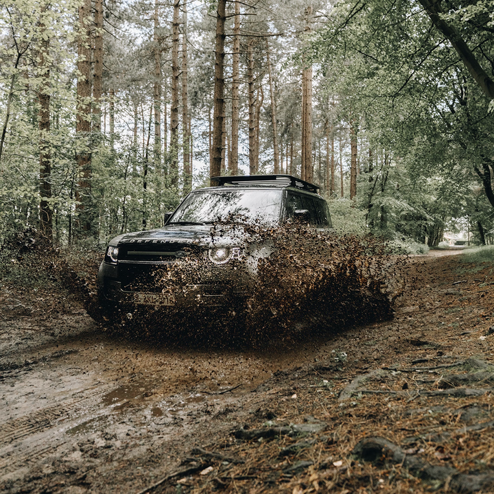 Black Land Rover Defender with a roof rack driving through deep mud in a forest
