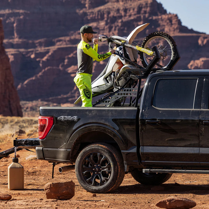 Man loading dirt bike onto black 4x4 truck in desert with red rock cliffs, overlanding setup