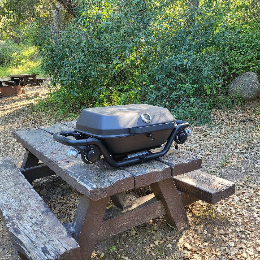 Portable grill on a wooden picnic table outdoors with greenery in the background