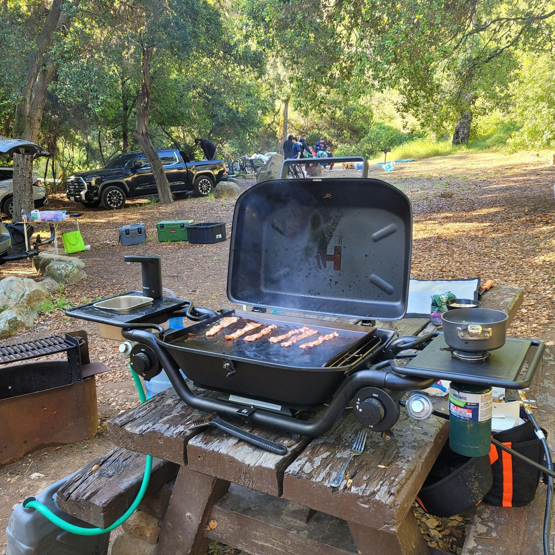 Portable grill on a picnic table with food being cooked, surrounded by trees and camping equipment.