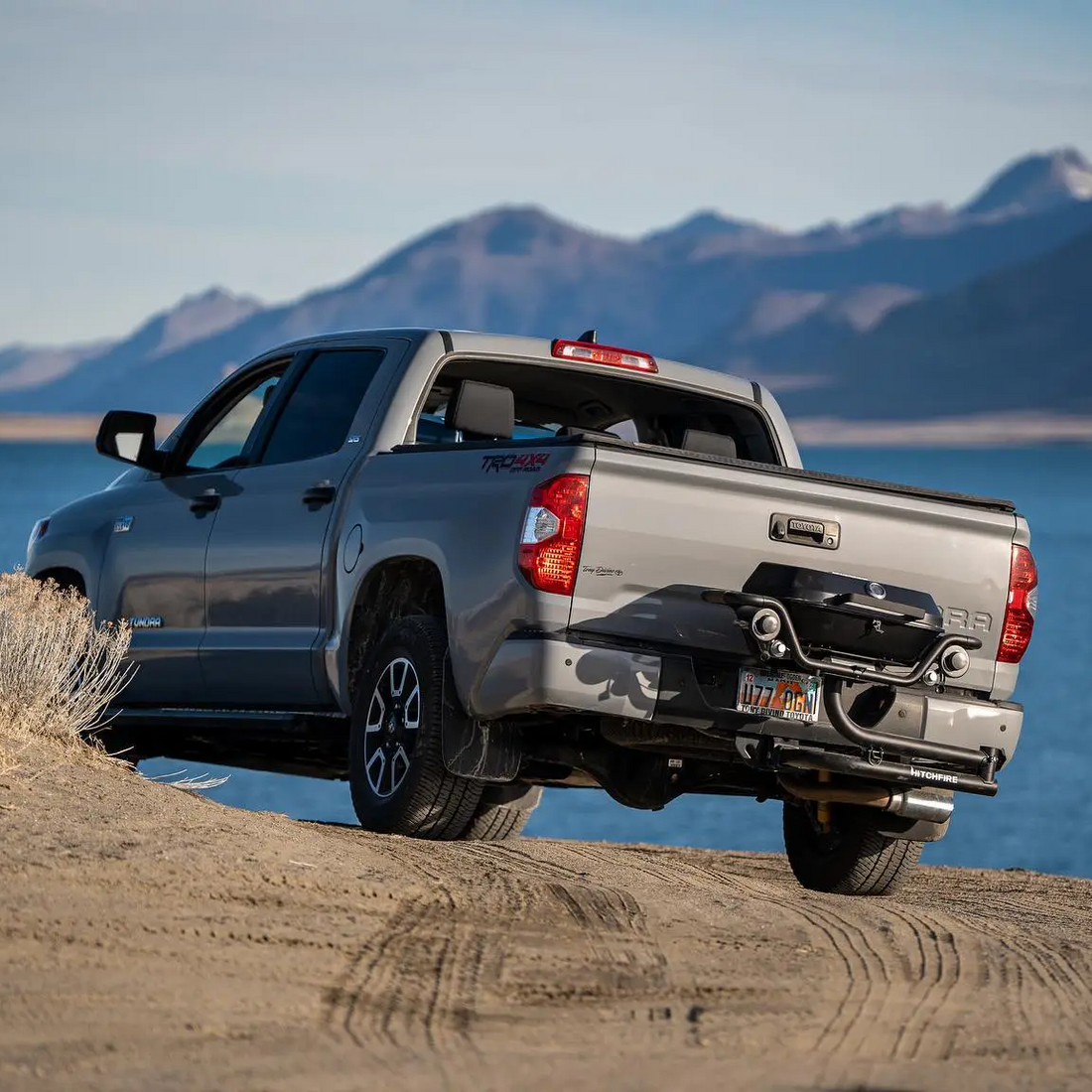 Silver pickup truck on a dirt road with mountains and water in the background