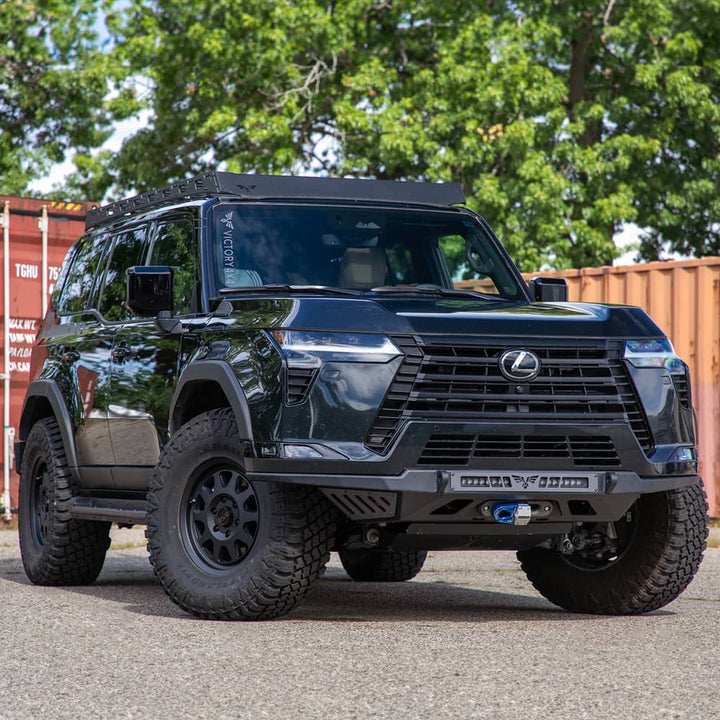 A black Lexus SUV with off-road modifications, including a front bumper guard and roof rack, parked outdoors in front of a rusted shipping container with trees in the background.