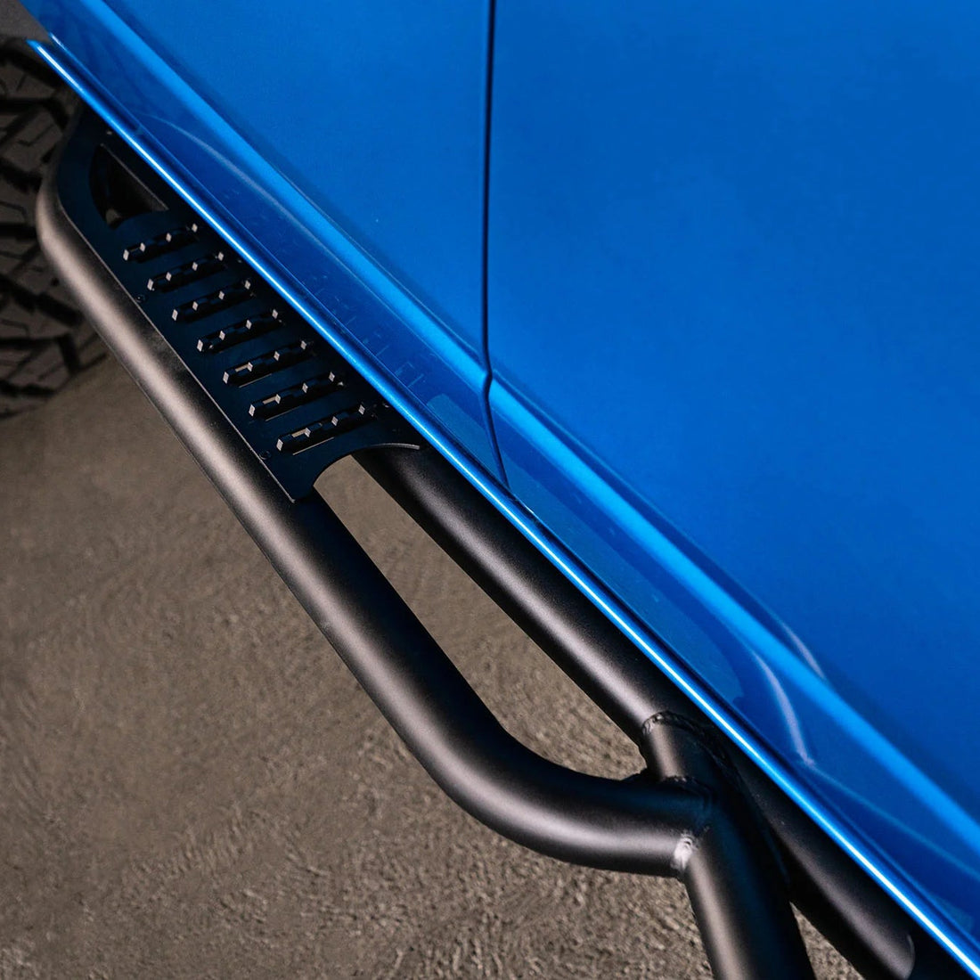 Close-up of a blue vehicle with a black side step bar on a textured surface.