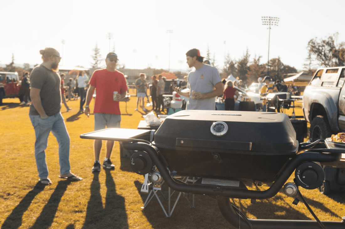 People standing around a grill at an outdoor event with cars and people in the background.
