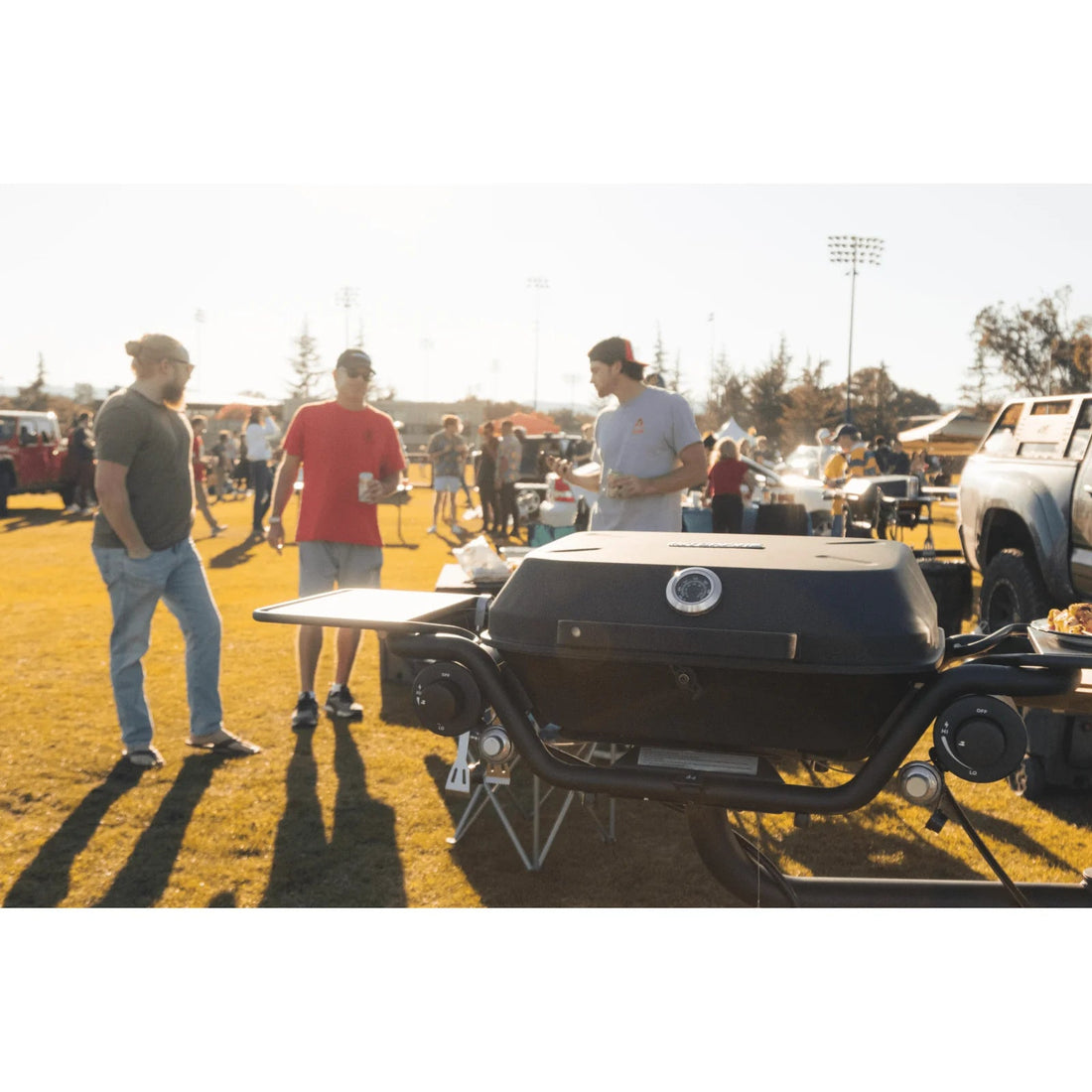 People standing around a grill at an outdoor event with cars and people in the background.