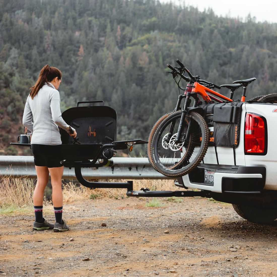 Person preparing a grill on a truck bed with bicycles attached, set against a forest backdrop.