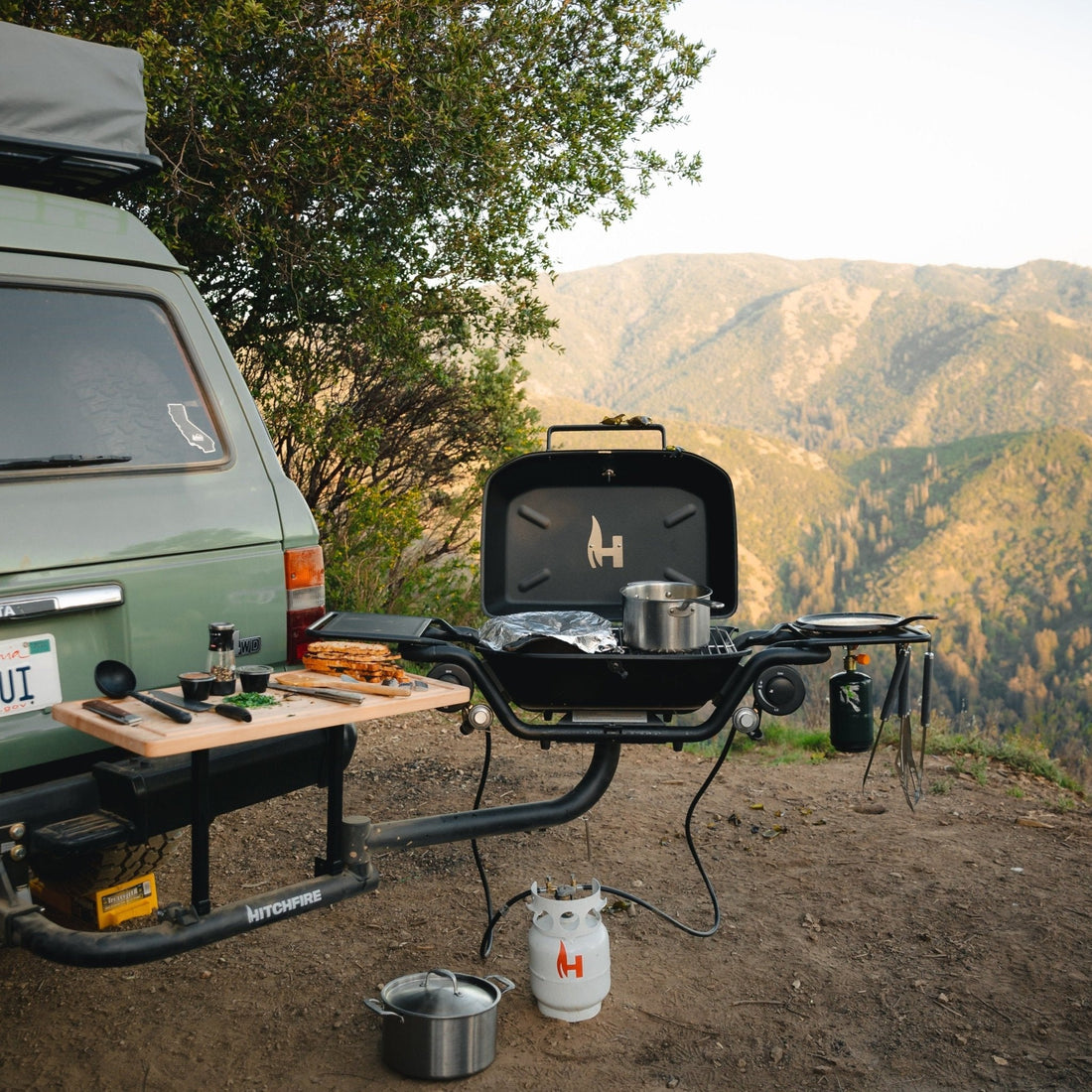 Outdoor camping setup with a grill and cookware near a vehicle in a mountainous area.