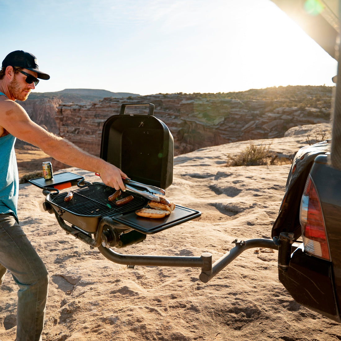 Man grilling outdoors in a desert landscape with a vehicle and scenic view.